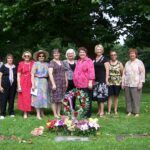 group of smiling women around a grave marker wreath and flowers